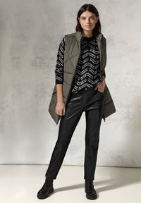 Olijfgroen gewatteerd vest over een zwart-wit zigzagpatroon sweater, gecombineerd met zwarte leren broeken en zwarte veterschoenen.