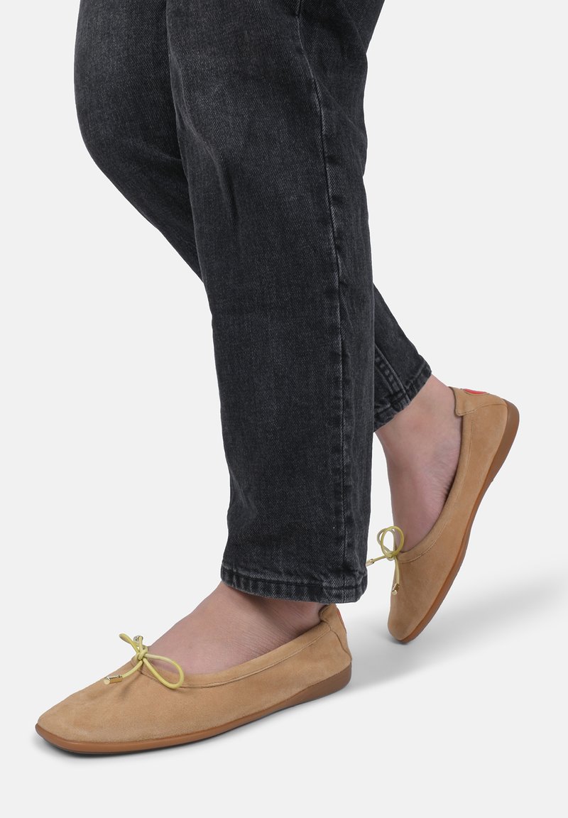 Ballerines en suede beige avec un bout arrondi et un petit accent de nœud jaune, associées à un jeans noir. La semelle en caoutchouc plat offre de la stabilité.