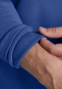 Pullover blu con un esterno texturizzato e polsini foderati in morbido pile. Primo piano di una mano che regola il polsino, evidenziando lo spessore del tessuto.