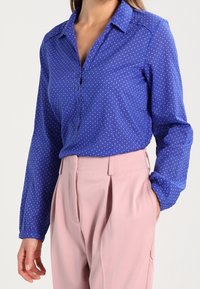 Camicia azzurra con bottoni, a pois bianchi, maniche lunghe e colletto, abbinata a pantaloni rosa chiaro a vita alta.