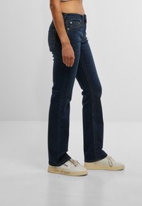 O persoană stă de profil, purtând blugi negri și adidași albi, punând în evidență o ținută casual și stilată pe un fundal simplu.