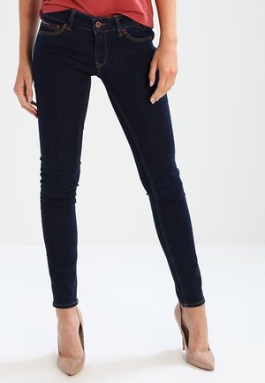 Femme portant un jean skinny bleu foncé, un haut rouge et des chaussures à talons hauts beige, debout devant un fond clair uni.