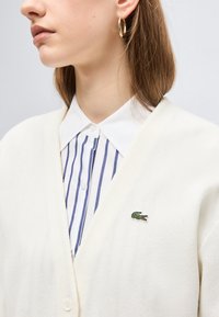 Femme portant un cardigan blanc avec un logo crocodile vert sur une chemise boutonnée à rayures bleues et une boucle d'oreille dorée en forme d'anneau, regardant vers la gauche.