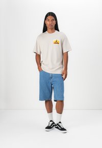 Beige bomull t-skjorte med et lite grafisk trykk, kombinert med blå denimshorts. Svart-hvite sneakers og hvite sokker fullfører antrekket.
