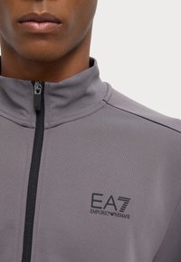 Giacca sportiva grigia con colletto alto e zip nera, caratterizzata dal logo EA7 Emporio Armani sul petto e da un tessuto liscio e texturizzato.