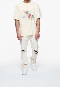 T-shirt color crema con grafica frontale di bevande, abbinata a jeans bianchi strappati con buchi alle ginocchia e sneakers bianche.