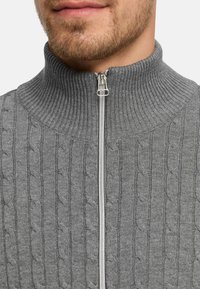 Grå kabelstrikket genser med høy krage, hel glidelås foran og ribbestruktur. Nærbilde fremhever det intrikate mønsteret og sømmene.