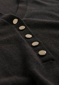 Gros plan d'un tissu tricoté noir avec cinq boutons ronds argentés brillants alignés verticalement sur une patte de boutonnage.