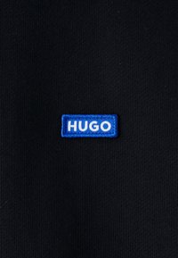 Etiqueta de tela rectangular azul con texto blanco "HUGO" cosida sobre un fondo de tela negra.