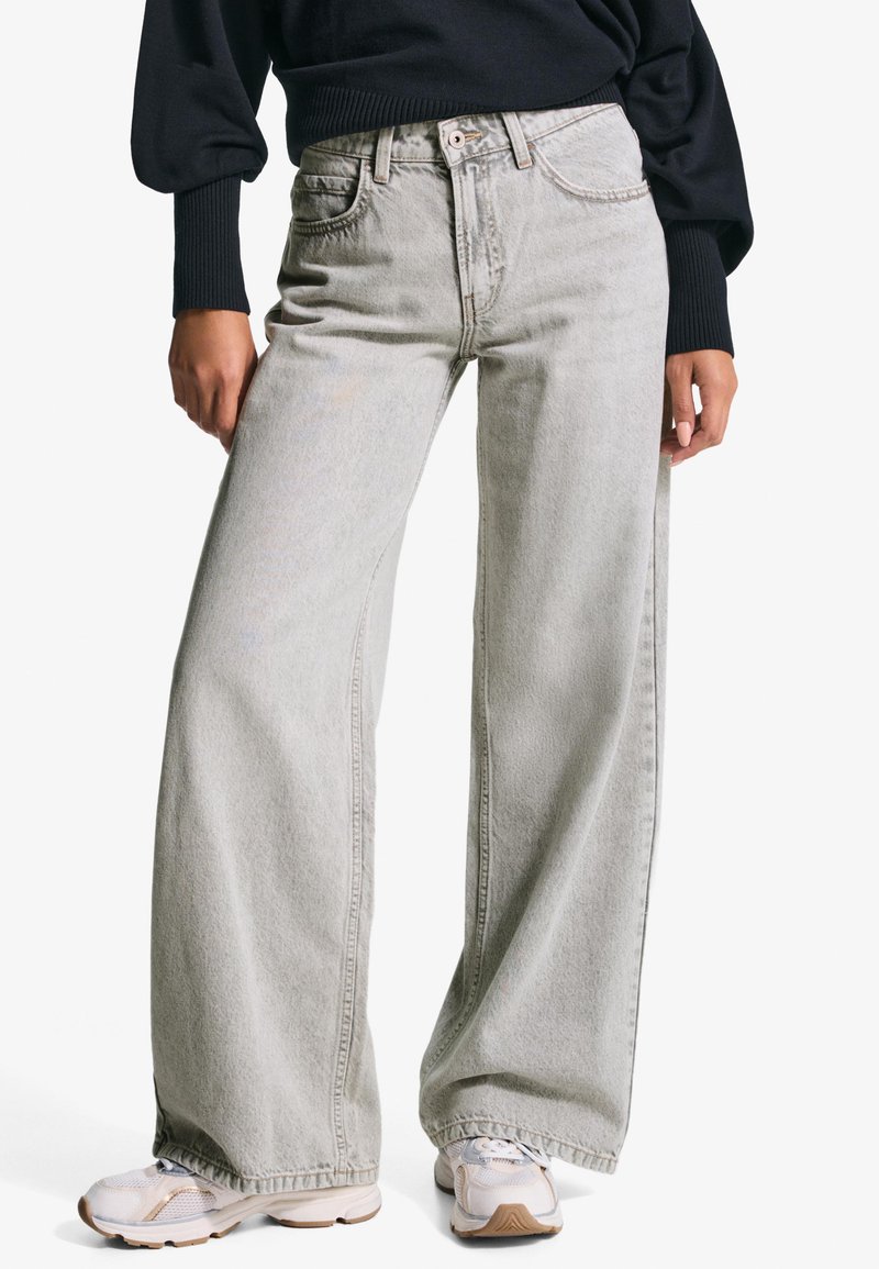 Világosszürke, bő szabású farmernadrág, magas derékkal, elülső zsebekkel és enyhén kopott hatással, fehér és bézs sportcipővel párosítva.