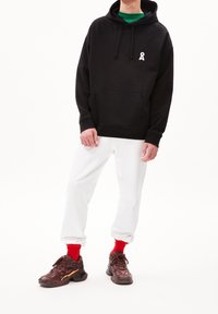Felpa nera con cappuccio e tasca anteriore, contrassegnata da un logo bianco, abbinata a pantaloni della tuta bianchi e sneakers marroni chunky con accenti rossi.