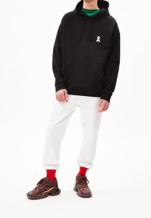Svart huvtröja med framficka och vitt logotyp, tillsammans med vita träningsbyxor och kraftiga bruna sneakers med röda detaljer.