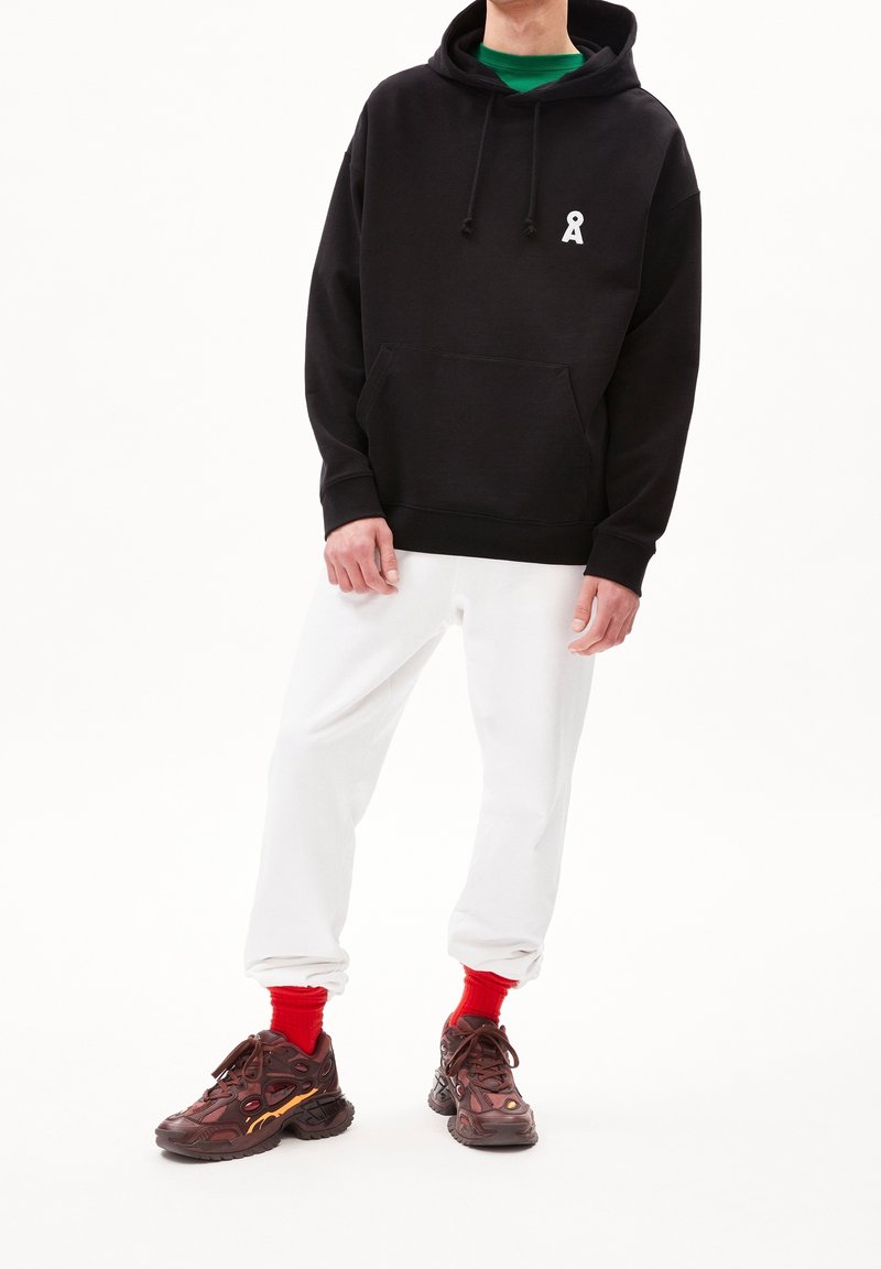 Felpa nera con cappuccio e tasca anteriore, contrassegnata da un logo bianco, abbinata a pantaloni della tuta bianchi e sneakers marroni chunky con accenti rossi.