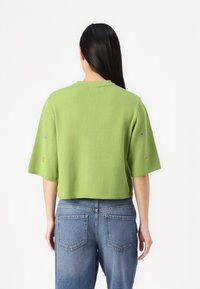 Grønn, kortermet strikket topp med rund hals og cropped design; har små blomsterbroderi på ermene og myk tekstur.