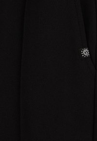 Fekete textúrájú szövet, egy kis fekete címkével, amelyen egy fehér spirális napdesign látható, és a varrás közelében van rögzítve.