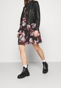 Veste en cuir noire portée sur une robe fleurie avec un fond noir et des fleurs roses. Bottes noires épaisses avec semelles épaisses et chaussettes blanches.