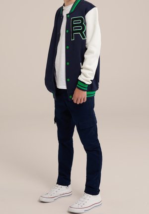Giacca varsity blu navy con maniche bianche e rifiniture verdi. Presenta una grande lettera "R" verde e bottoni sul davanti. Abbinata a pantaloni cargo e scarpe da ginnastica.