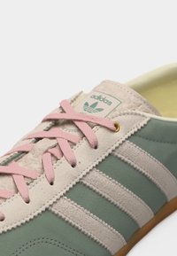 Κοντινό πλάνο ανοιχτοπράσινου αθλητικού παπουτσιού Adidas με λευκές ρίγες και ροζ κορδόνια, που δείχνει το λογότυπο στη γλώσσα και υφασμένα σουέντ λεπτομέρειες.