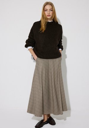 Jeune femme debout, les mains dans les poches, portant un pull marron foncé, les poignets d'une chemise blanche visibles, une longue jupe beige à motifs et des mocassins noirs.