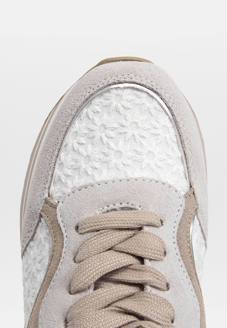 Sneaker grigio chiaro con tomaia in suede, ricami floreali bianchi e un pannello in metallo argentato. Lacci rotondi beige e suola testurizzata.