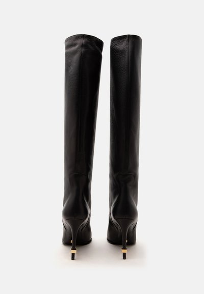 Paire de bottes hautes en cuir noir à talons fins montrée de dos avec des accents dorés près des semelles sur un fond uni.