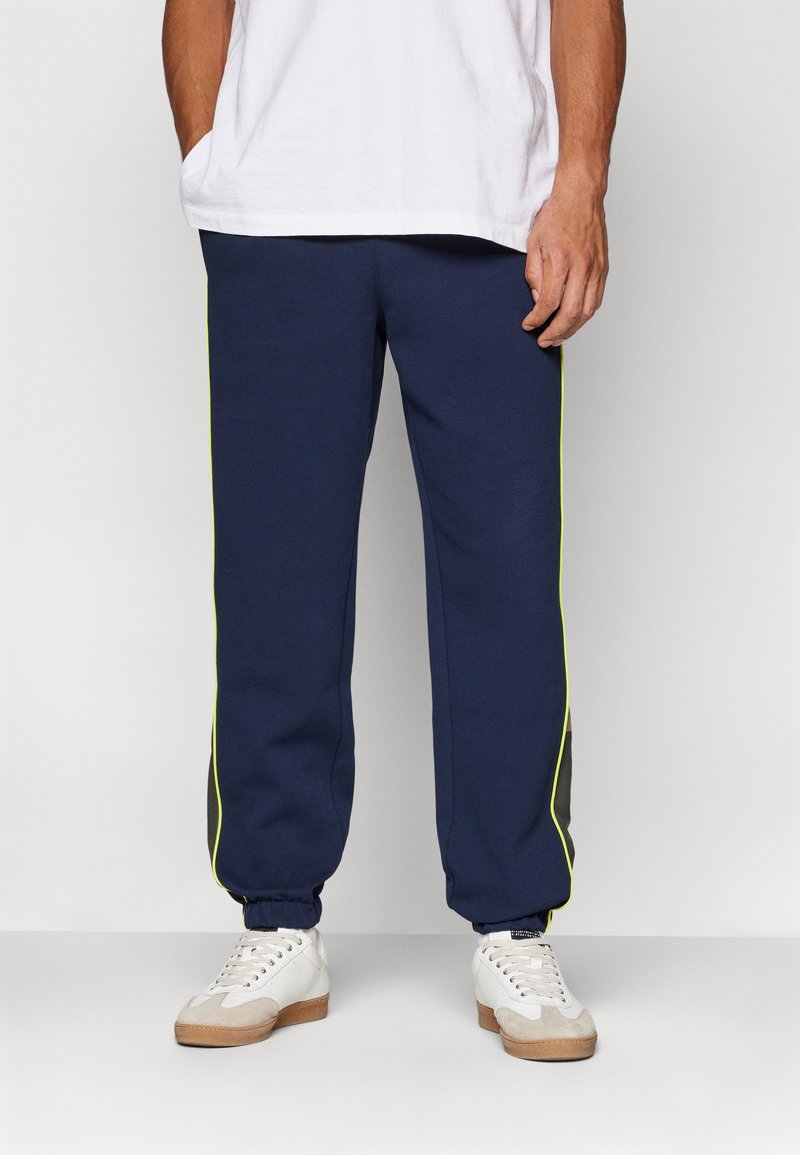 Calças de felpa azul-marinho com punhos elásticos, riscas laterais em amarelo neon contrastante e uma textura suave, combinadas com ténis brancos e uma t-shirt branca.