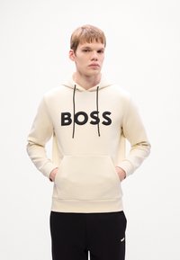 Jaunas vyras dėvi kreminės spalvos megztinį su užrašu "BOSS" ir juodas kelnes, rankos kišenėje, stovi prieš baltą fono.