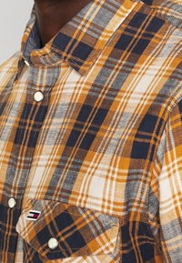 Primo piano di una camicia a quadri gialla senape, blu navy e bianca, con tasca a pattina con bottoni, e bottoni a pressione bianchi sul colletto e nella parte frontale.