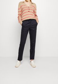 Beżowy sweter z dzianiny w czerwone paski, z dekoltem w serek i kołnierzykiem; granatowe, zwężające się spodnie; białe trampki z fakturowaną podeszwą.