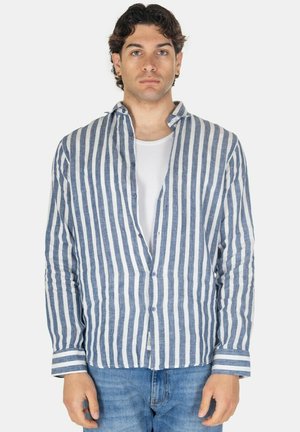 Giovane uomo con capelli corti e scuri che indossa una camicia a righe verticali blu e bianche sopra una t-shirt bianca e jeans blu, in piedi dritto.