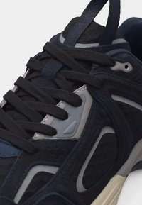 Κοντινή λήψη ενός αθλητικού παπουτσιού σε ναυτικό μπλε και μαύρο με παχιά μαύρα κορδόνια, πάνελ από δίχτυ και συνθετικά υλικά, και λεπτομέρειες στη σόλα σε μπεζ χρώμα.