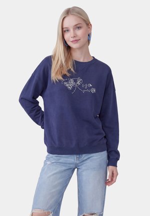 Ung kvinde med langt blondt hår iført blå hoops-øreringe, en navy sweatshirt med rhinsten sommerfugl og rosedesign og revnede lyseblå jeans.