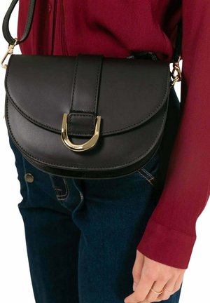 Sac bandoulière en cuir noir avec boucle dorée porté sur une chemise rouge à manches longues et un jean bleu foncé, main détendue le long du corps.