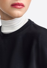 Svart sweatshirt med en slät textur och en rundad halsdesign, lagd över en vit polotröja. Närbild på halsringningen och nedre delen av ansiktet.