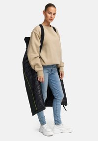 Schwarze Pufferjacke mit einer gesteppten Textur und neon-grünen Akzenten, getragen über einem beigen Sweatshirt und hellblauen Jeans, kombiniert mit weißen Sneakers.