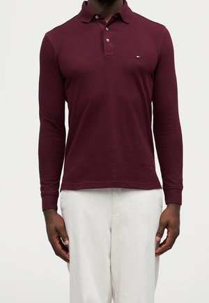 Mężczyzna ubrany w bordową koszulkę polo z długim rękawem i guzikami, w zestawie z jasnobeżowymi spodniami, stojący na jednolitym tle.