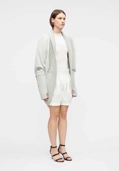 Γυναίκα που στέκεται φορώντας μια γκρι υπερμεγέθη ζακέτα, λευκό πουκάμισο, λευκά σορτς με σούρες και μαύρα ψηλοτάκουνα σανδάλια με λουράκια, μπροστά σε λευκό φόντο.