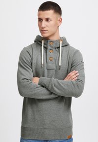 Sudadera con capucha gris y cuello alto, botones decorativos y cordones ajustables. Presenta un tejido texturizado y un parche con el logo en la manga.