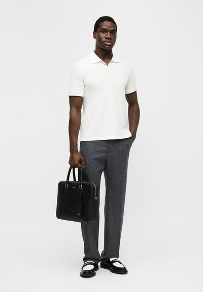 Homme portant un polo blanc à manches courtes, pantalon gris, mocassins noirs et blancs, tenant une mallette en cuir noir, debout devant un fond uni.