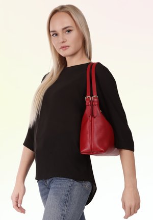 Blonde Frau mit schwarzem Oberteil und blauer Jeans, die eine rote Lederhandtasche auf der rechten Schulter trägt, vor hellem Hintergrund.