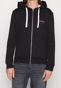 Persona vestida con una sudadera con cremallera negra y cordones blancos, y un logo circular rojo en el lado izquierdo del pecho, combinada con unos jeans gris oscuro.