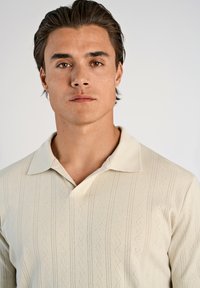 Ljusbeige stickad polo med ett texturerat mönster, som har en klassisk krage och en smal passform.