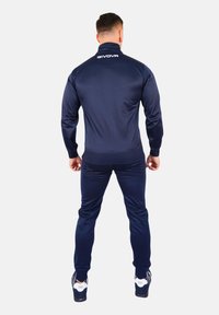 Tuta navy dintr-un material neted, cu un guler înalt și manșete ribbate. Logo-ul este afișat pe spate, având pantaloni tapered și adidași.