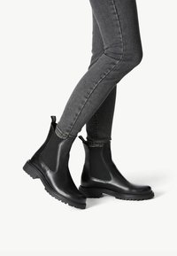 Botas Chelsea de cuero negro con paneles laterales elásticos, suelas de goma y tiradores. Combinadas con jeans ajustados grises, destacando su diseño elegante.