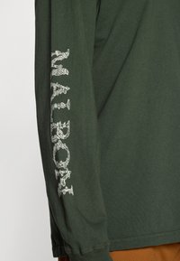 Chemise en coton vert olive à manches longues, ornée d'un design graphique blanc complexe le long de la manche gauche. Texture douce, coupe décontractée.