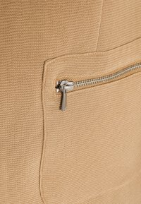 Beige texturerat tyg med en liten ficka med dragkedja och silverdetaljer. Fickan är omgiven av en subtil sömdetalj.