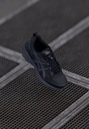 Sort ASICS sneaker med detaljeret syning og snørebånd, ophængt over et metalristgulv med krydsende linjer.