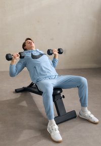 Világoskék sportpulóver nagy fekete logóval, hozzáillő leggings és fehér súlyemelő cipők. Fekete kézisúlyzókat tart a padon.