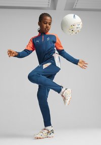 Chándal deportivo azul con acentos naranjas y patrones geométricos, combinado con zapatillas de fútbol de colores; se está pateando un balón de fútbol con habilidad.