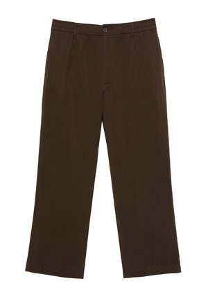 Personne debout, bras croisés, portant un t-shirt blanc, un pantalon marron foncé, une ceinture noire et des tongs noires devant un mur clair.
